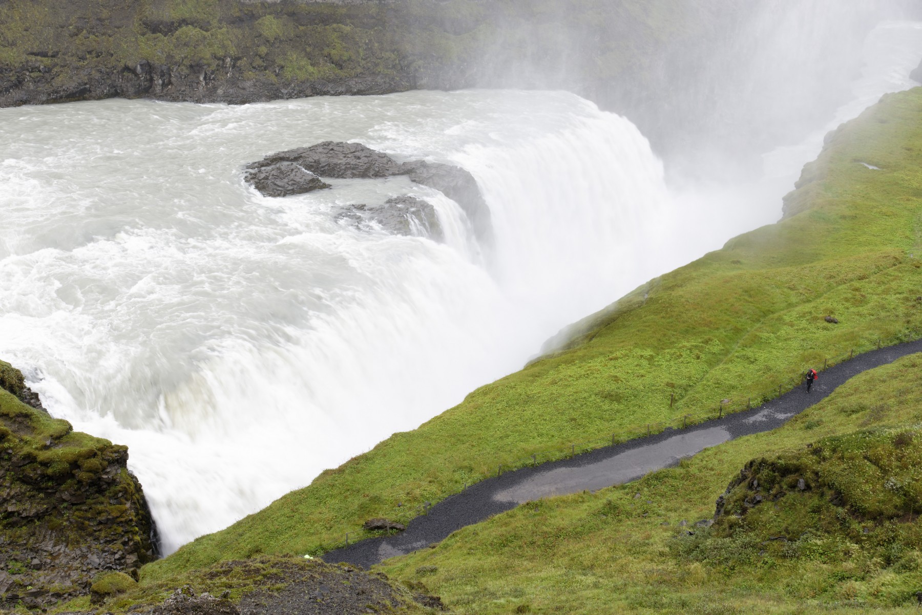 view from upper plattform at tourist on path parallel to gullfoss waterfall between lavarock wall and green grass field